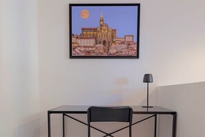 Ein gerahmtes Stadtbild an der Wand, ein Schreibtisch mit Stuhl und eine Stehlampe.