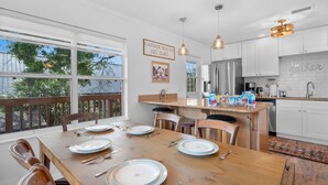 Un coin repas avec une table en bois dressée pour quatre personnes, une cuisine avec des meubles blancs et une vue sur la verdure à l’extérieur.