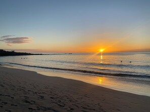 Ein Strand mit Fußspuren, die zum Wasser führen, ein Sonnenuntergang über dem Ozean und ein klarer Himmel.