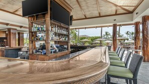 Un espace bar doté d’un comptoir incurvé, d’étagères en bois où sont exposées des bouteilles, et d’un coin salon extérieur agrémenté de parasols.