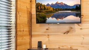 Puinen mökki sisältä, seinälle asennettu televisio, jossa näkyy vuoristojärvimaisema, puinen pöytä, jonka päällä on kynttilä, ja tumma puinen kaappi.