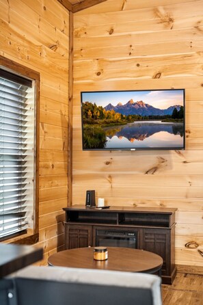 Interior de una cabaña de madera con un televisor montado que muestra una escena de un lago de montaña, una mesa de madera con una vela y un mueble de madera oscuro.