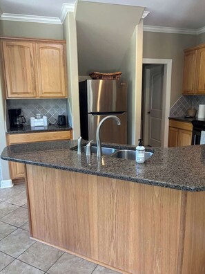 A kitchen with a granite countertop, stainless steel refrigerator, wooden cabinets, and a built-in sink. - Condo Townhome - F4 - 3 Bedrooms - Hot Springs (Garland County)