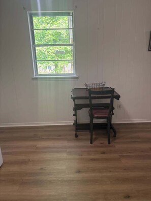 Una habitación con piso de madera, una ventana con vistas a la vegetación y un escritorio con una silla.