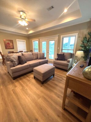 Ein Wohnzimmer mit Ecksofa, Hocker, Holzboden, Deckenventilator und großen Fenstern.