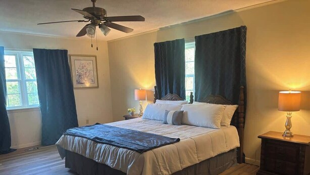 A bedroom with a bed, two bedside tables with lamps, a ceiling fan, and a window with curtains. - Large 4 Bedroom Home - Bryant, AR (Bryant)