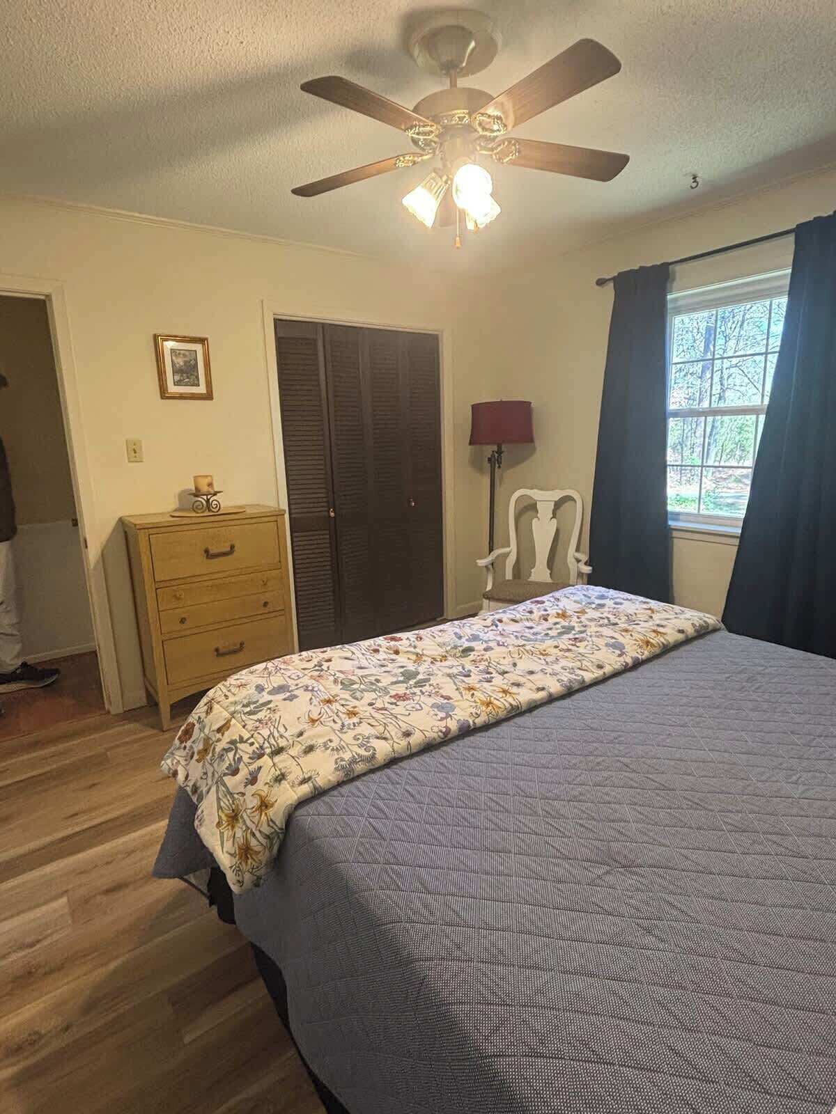 A bedroom with a bed, a ceiling fan, a dresser, a chair, a window with curtains, and a door.