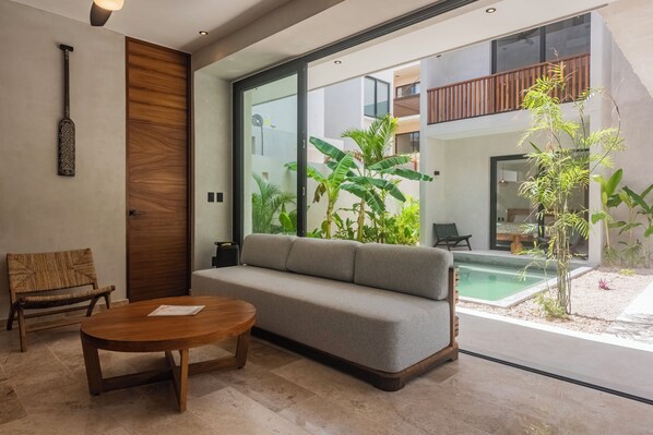 Ein modernes Wohnzimmer mit einer Couch, einem Holztisch, einem Korbstuhl und einer großen Glasdoor, die zu einem Poolbereich führt.