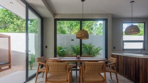 Un espace repas moderne avec un mur en verre, des chaises en osier, une table en bois et un luminaire suspendu.
