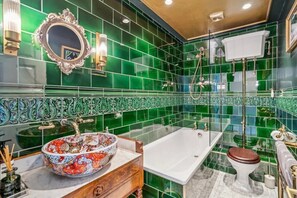 A bathroom with green tiles, a white bathtub, a wooden vanity, a colorful sink, and a mirror. - Luxury Kensington Townhouse near Hyde Park (London)