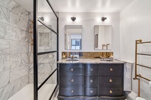 Uma casa de banho moderna com azulejos de mármore, toucador preto com detalhes dourados e um espelho grande.
