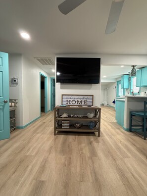 A modern living room with a flat-screen TV, a wooden console table with decorative items, teal-colored walls, and a kitchen area in the background. - Hidden Oasis- 2 bdrm 2 bath home (Port O'Connor)