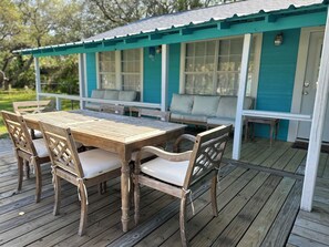 En træterrasse med et bord og stole, et overdækket siddeområde med hynder, og en teal farvet facade.