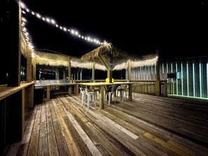 Un espace bar extérieur doté d’un plancher en bois, de parasols à toits de chaume et de guirlandes lumineuses.