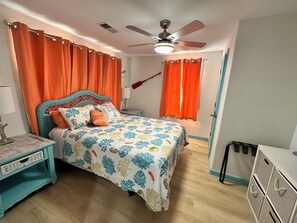 Ein Schlafzimmer mit Bett, Nachttisch, Deckenventilator und einem Fenster mit Vorhängen.