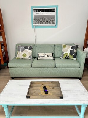 Ein grünes Sofa mit gemusterten Kissen, eine Fernbedienung auf einem Holztisch und eine Wand-Klimaanlage.