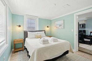 Ett sovrum med en säng, sängbord, ett fönster med persienner, en tavla på väggen och ett litet kök i det intilliggande rummet.