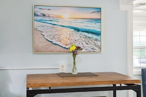 Ein gerahmtes Strandbild an einer Wand, ein Holztisch mit Blumen in einer Vase und eine Steckdose.