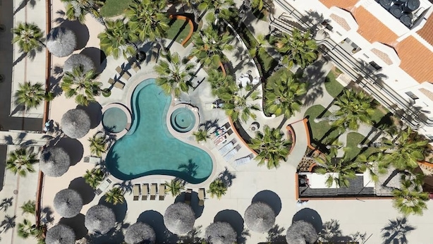 Vue aérienne d’un complexe hôtelier doté d’une piscine centrale, entourée de palmiers et d’espaces de détente.