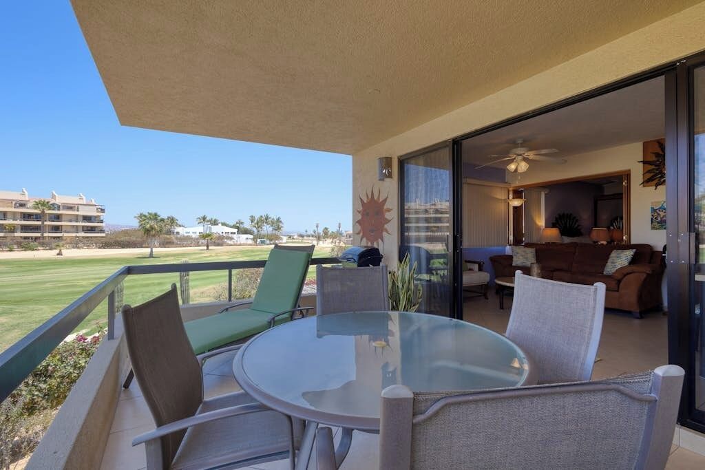 A balcony with a glass table and chairs, a view of a golf course, and a glimpse of the interior with a couch and a ceiling fan.