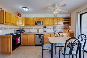 A kitchen with wooden cabinets, a microwave, a dishwasher, a stove, and a dining area with chairs and a table. - Charming Golf-view Condo With Pool, Hot Tub, Terrace & Wifi (San José del Cabo)