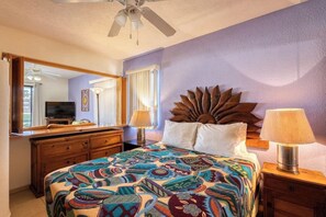 Une chambre à coucher avec un lit, un ventilateur de plafond, une commode, un téléviseur et une fenêtre avec des rideaux.