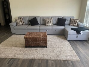 Un canapé d’angle gris avec des coussins à motifs, un pouf en osier et un tapis de couleur claire dans un salon doté d’un plancher en bois.