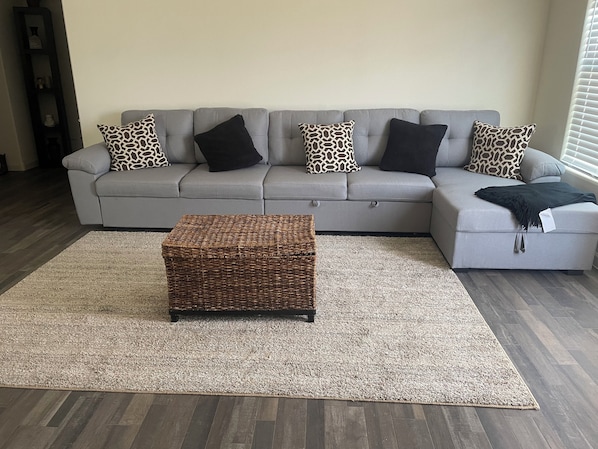 Um sofá modular cinza com almofadas estampadas, um banquinho trançado e um tapete em tons claros em uma sala de estar com piso de madeira.