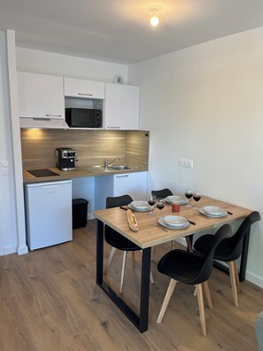 Una cocina funcional con microondas, tostadora y cafetera, una mesa de comedor con platos y vasos, y un refrigerador pequeño.