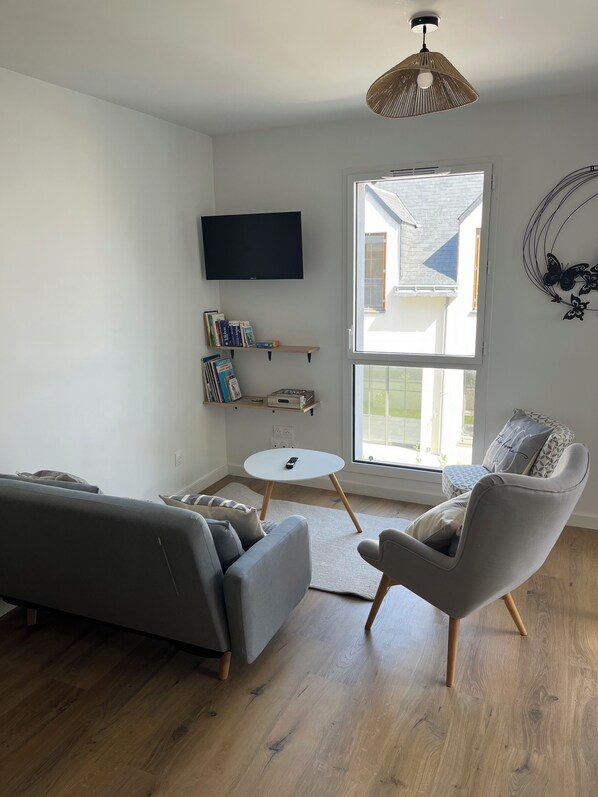 Una sala de estar moderna con un sofá gris, un sillón gris, una mesita blanca y un televisor montado en la pared. Hay estanterías debajo del televisor y una ventana con vistas al exterior.