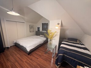 Ein Schlafzimmer mit schrägem Dach, einem Bett mit gestreifter Bettdecke, einem Nachttisch mit Blumen in einer Vase, einer Wandlampe und einer Tür.