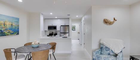 A modern kitchen with white cabinets, a dining area with wicker chairs, and a blue patterned armchair.