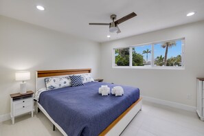 Ein Schlafzimmer mit einem Bett, einem Nachttisch, einem Deckenventilator, einem Fenster mit Blick auf Palmen und einer weißen Wand.