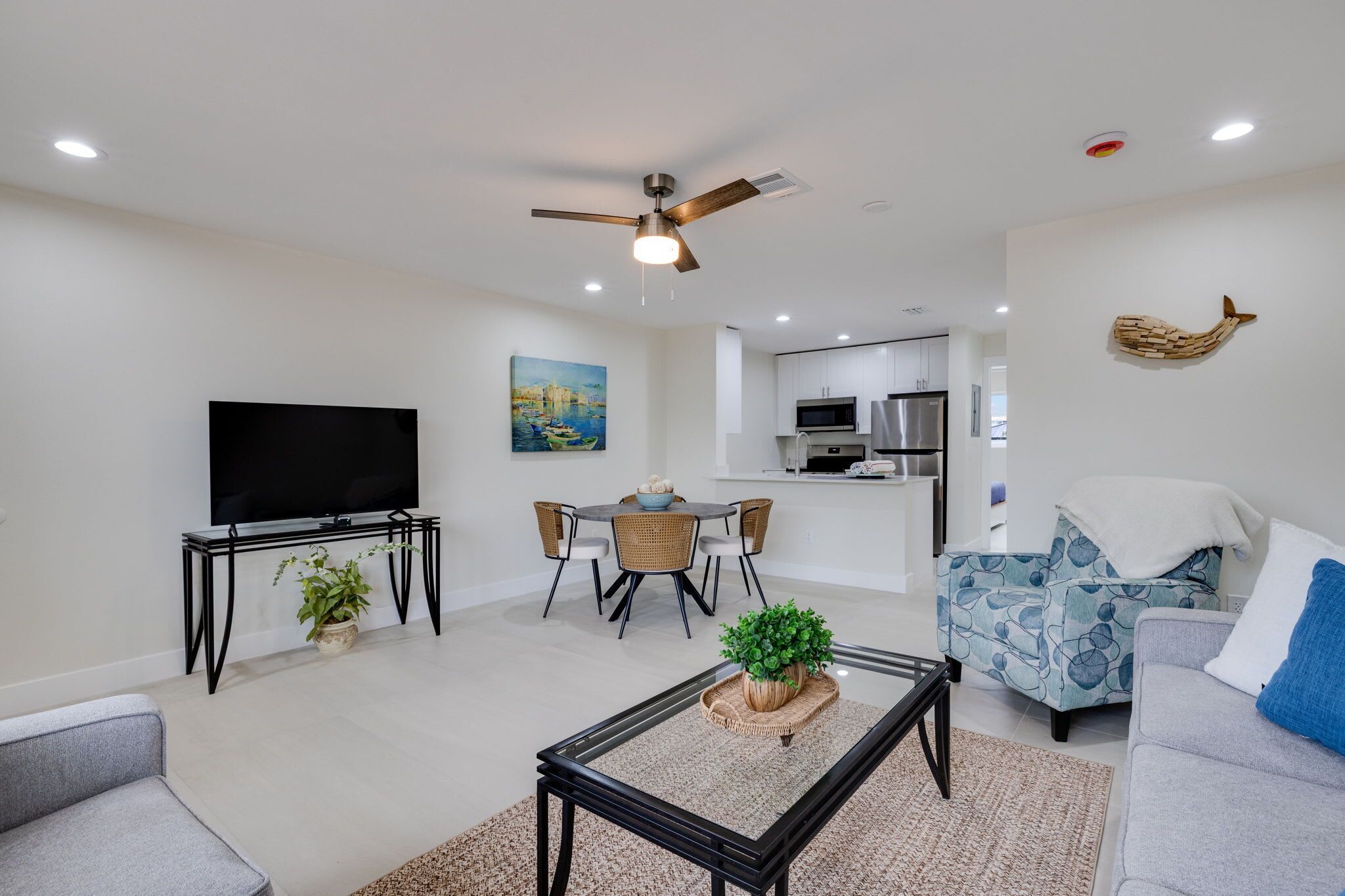 A living room with a TV, a ceiling fan, a dining area with chairs, a kitchen in the background, and a sofa with a cushion.