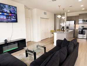 Una sala de estar moderna con televisor de pantalla plana, una mesa de centro de vidrio, un sofá negro y una zona de cocina con electrodomésticos de acero inoxidable.