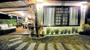 A covered outdoor seating area with a sofa and tables, a window with curtains, and a garden with plants and stepping stones. - Jardim do Mar - Beachfront Pool, Sports Area, Resort Style (Armação dos Búzios)