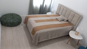 Une chambre à coucher moderne avec un lit, un fauteuil en forme de poire, une petite table blanche et une fenêtre avec des rideaux légers.