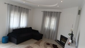 Un salon avec un canapé noir, un pouf bleu, une télévision et une petite plante.