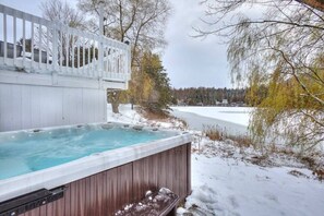 Ein Whirlpool mit Holzterrasse und weißer Reling, umgeben von Schnee und Bäumen.