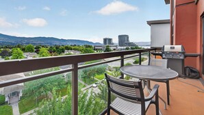 A balcony with a grill, a table, and chairs, overlooking a cityscape with buildings and greenery. - Playa Del Sol 2 Bd 649 w Mountain Views (Kelowna)