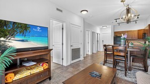 A living room with a flat-screen TV, a wooden coffee table, and a dining area with a kitchen in the background. - Playa Del Sol 2 Bd 649 w Mountain Views (Kelowna)