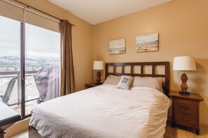 Quarto de hotel com uma cama grande, duas mesas de cabeceira com luminárias, uma janela com cortinas e dois quadros pendurados na parede.