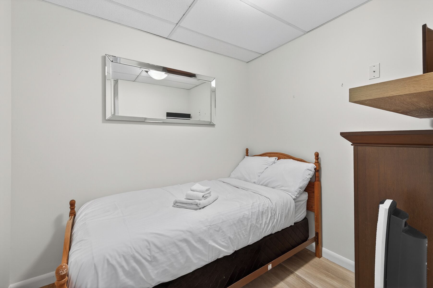 A single bed with a wooden headboard, a white mattress, and a neatly folded towel. A mirror hangs on the wall above the bed, and a wooden shelf is mounted above the bed.