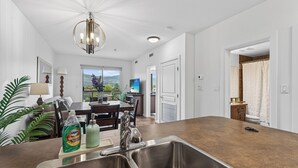A modern kitchen with a central island, stainless steel sinks, and a dining area with a view. - Playa Del Sol 2 Bd 649 w Mountain Views (Kelowna)