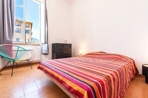 A bedroom with a bed, a chair, a radiator, and a window with a view of buildings. - Casa Soprana - À moins d'1 km de la plage (L'Île-Rousse)
