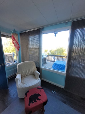 A room with a chair, a small table with a bear design, a window with a view of a deck, and a ceiling fan.