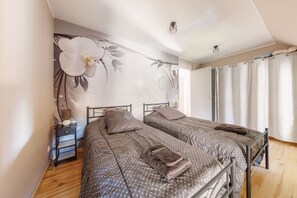 A bedroom with two beds, a nightstand, a wall with floral wallpaper, and a window with curtains. - Maison Paulette - Belle maison pour 8 en altitude (Neussargues en Pinatelle)