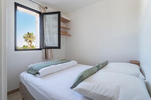 A bedroom with a bed, a window with a view of palm trees, a hanging laundry rack, and a bedside table. - Les Capitelles - Avec accès direct à la plage (Saint-Cyprien)