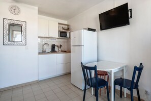 A compact kitchen with white cabinets, a microwave, a refrigerator, a dining table with two chairs, and a wall-mounted TV. - Les Capitelles - Avec accès direct à la plage (Saint-Cyprien)