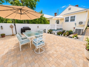 Een terras met een tafel, stoelen, een parasol, een barbecue en een witte hek.
